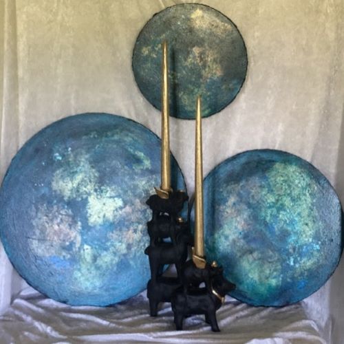 Tre runde malerier i blått og turkis med gullmetallisk overflate, fotografert med sorte lysestaker og gullstaker foran hvit bakgrunn. 3 univers av Art by Emerense
