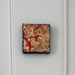 Nærbilde av kvadratisk kunstverk med gullblader og røde penselstrøk på sort kant. Mix media, 10×10 cm. Art by Emerense