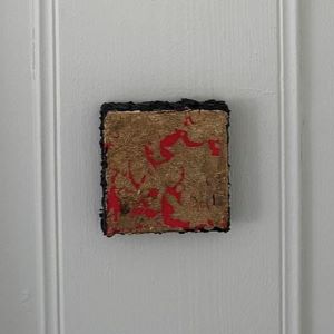 Kvadratisk kunstverk med gylden overflate og røde detaljer i mix media. Sort kant, 10×10 cm. Art by Emerense