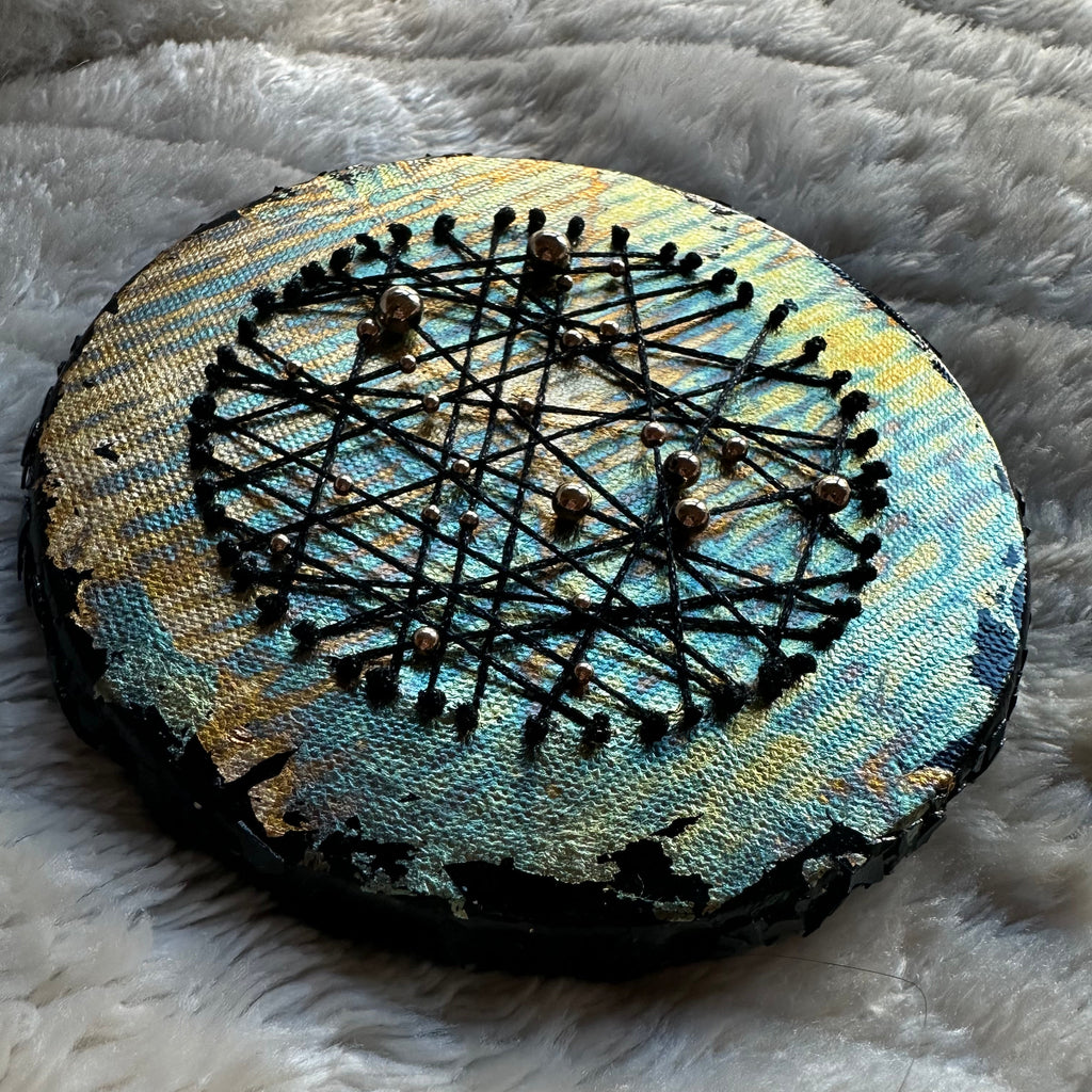 Constellations IV, rundt mixed media-kunstverk med metalliske gull- og turkistoner, kryssende svarte tråder og små gullfargede detaljer, fotografert på et lyst, mykt underlag.