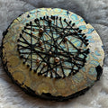 Constellations VI, rundt mixed media-kunstverk i metalliske gull- og turkistoner med kryssende svarte tråder og små gullfargede detaljer, fotografert på et lyst, mykt underlag.