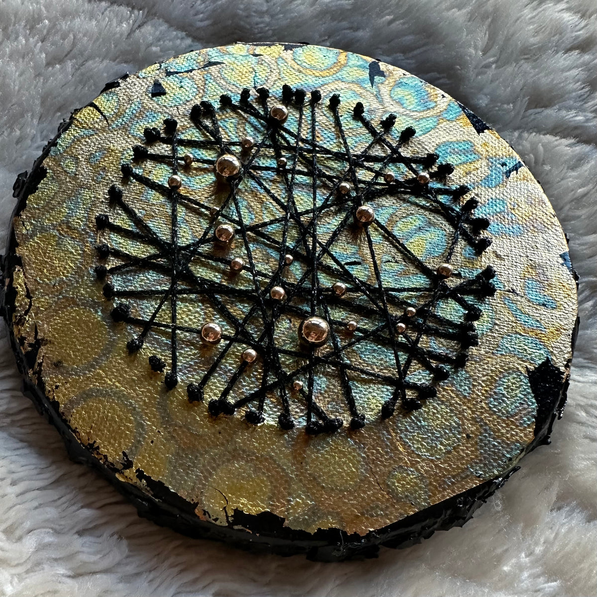 Constellations VI, rundt mixed media-kunstverk i metalliske gull- og turkistoner med kryssende svarte tråder og små gullfargede detaljer, fotografert på et lyst, mykt underlag.