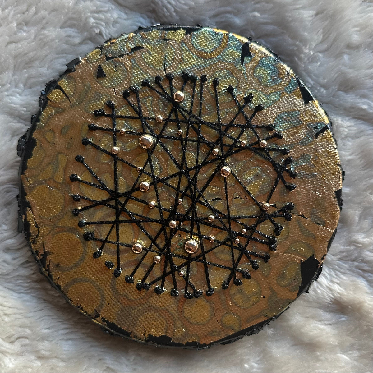 Constellations VI, rundt mixed media-kunstverk i metalliske gull- og turkistoner med kryssende svarte tråder og små gullfargede detaljer, fotografert på et lyst, mykt underlag.