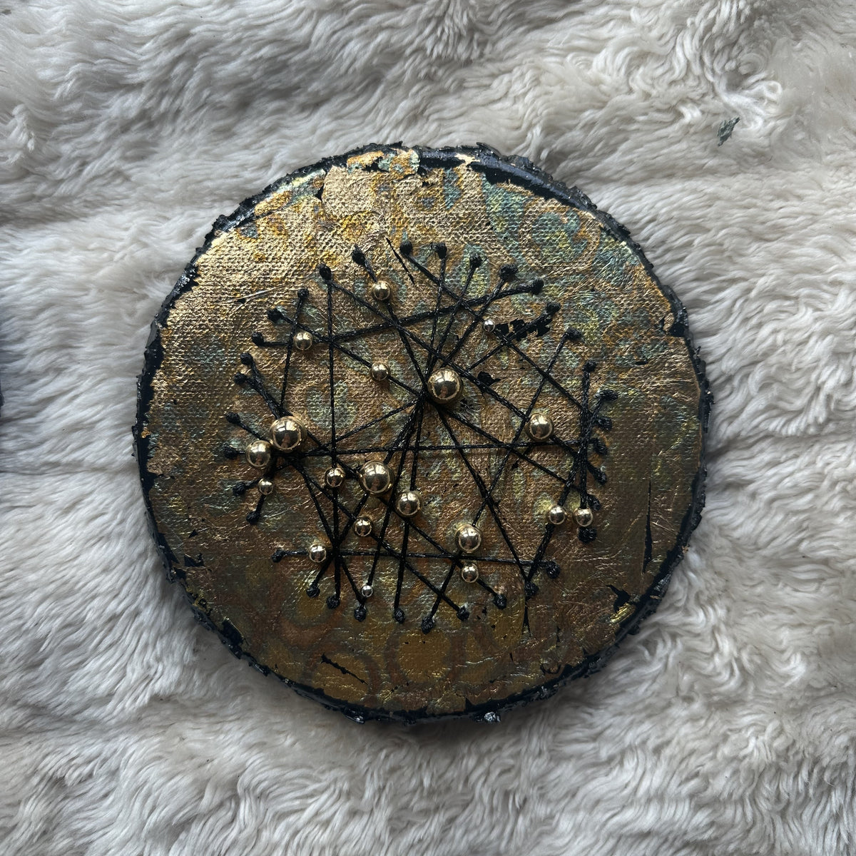 Rundt mixed media-kunstverk, 15 cm diameter, med gull og grønn-turkis iriserende overflate. Sorte tråder og gullperler i stjernemønster. Constellations III av Art by Emerense
