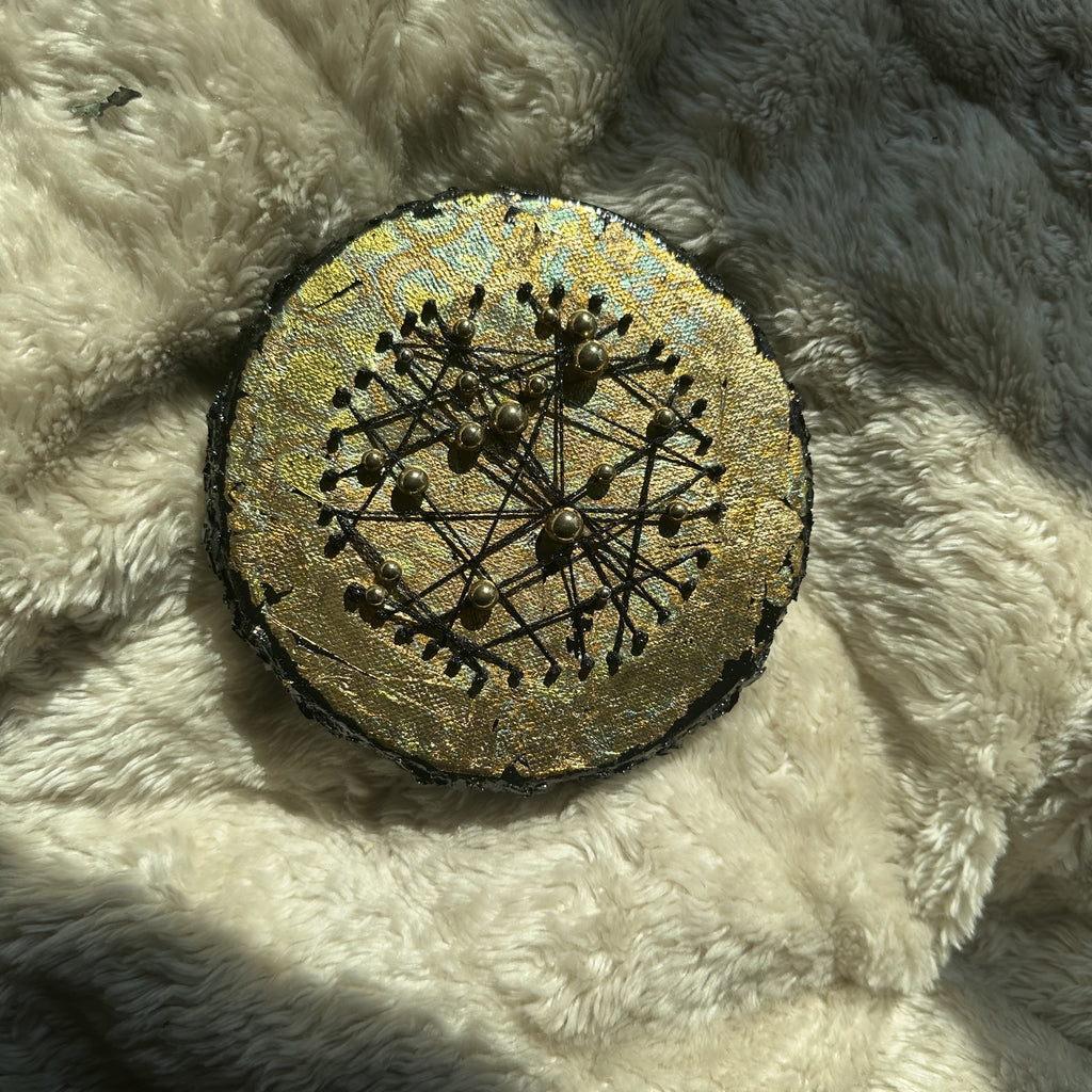 Rundt mixed media-kunstverk, 15 cm diameter, med gyllent skimrende overflate og sorte tråder med perler i konstellasjonsmønster. Constellations III av Art by Emerense