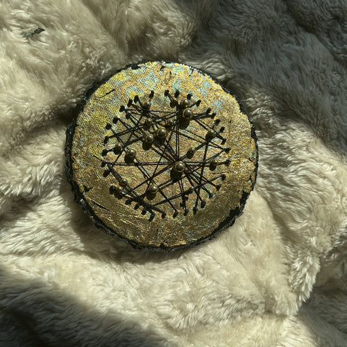 Rundt mixed media-kunstverk, 15 cm diameter, med gyllent skimrende overflate og sorte tråder med perler i konstellasjonsmønster. Constellations III av Art by Emerense