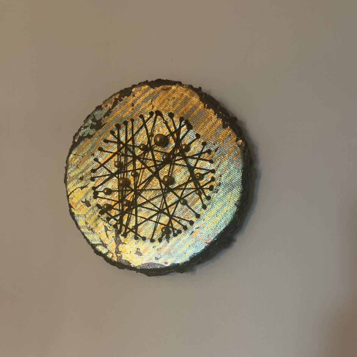 Rundt mixed media-kunstverk, 15 cm diameter, hengt på beige vegg. Sølv og gull iriserende overflate med sorte tråder og metallperler. Constellations III av Art by Emerense
