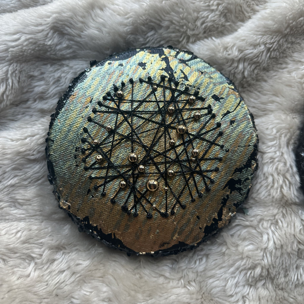 Rundt mixed media-kunstverk, 15 cm diameter, med sølv og blågrønn iriserende overflate. Sorte tråder og sølvperler i stjernemønster mot sort kant. Constellations I av Art by Emerens
