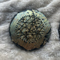 Rundt mixed media-kunstverk, 15 cm diameter, med sølv og blågrønn iriserende overflate. Sorte tråder og sølvperler i stjernemønster mot sort kant. Constellations I av Art by Emerens
