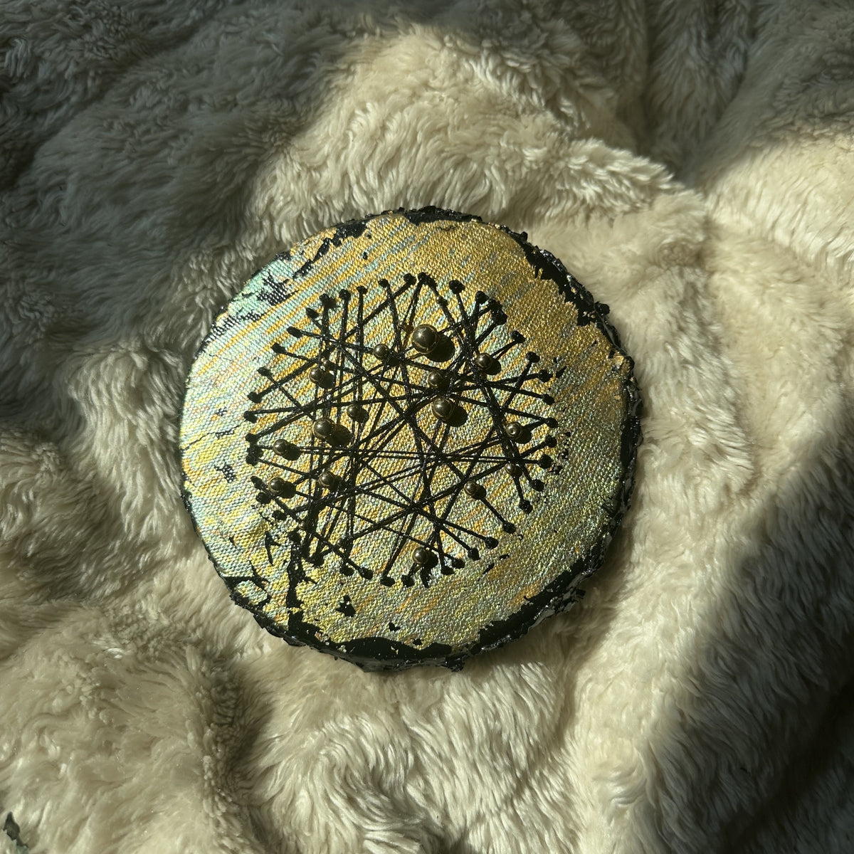 Rundt mixed media-kunstverk, 15 cm diameter, sølv og blågrønn skimrende overflate med sorte tråder og metallperler. Fotografert i naturlig lys på fluffy overflate. Constellations I av Art by Emerense
