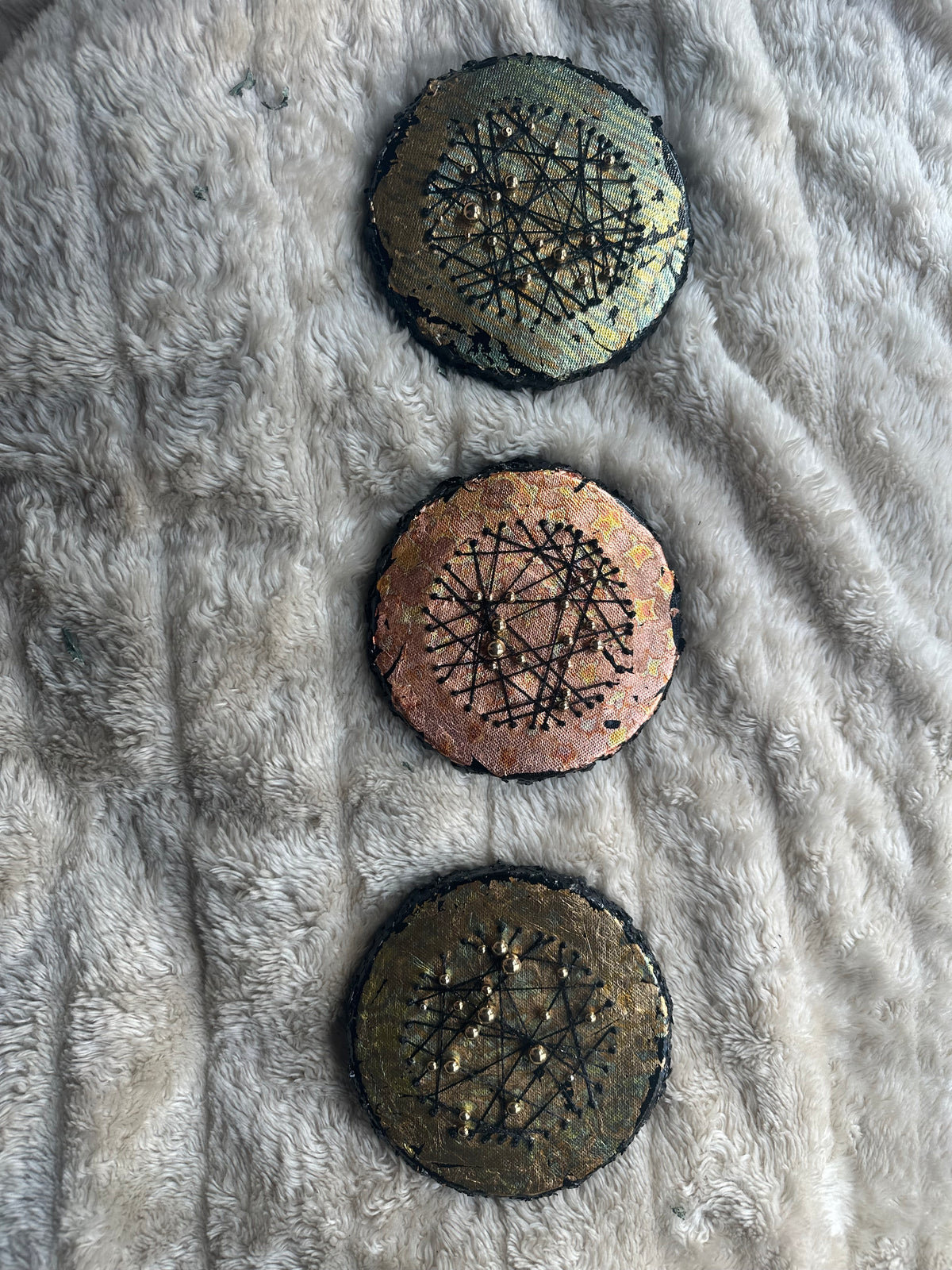 Tre runde mixed media-kunstverk, 15 cm i diameter, lagt vertikalt på grå fluffy overflate. Sorte tråder og metalliske perler i grønn, kobber og gull mot mørk bakgrunn. Constellations sett med 3 av Art by Emerense
