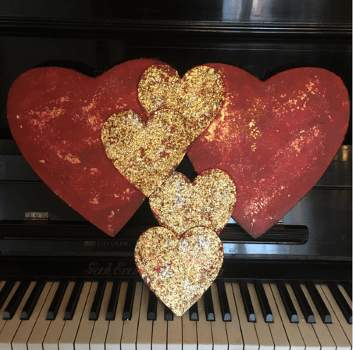 To store røde hjerter med gullflak og tre mindre gullhjerter i ulike størrelser, fotografert foran et sort piano. Familiekjærlighet, akryl og mix medium. Art by Emerense
