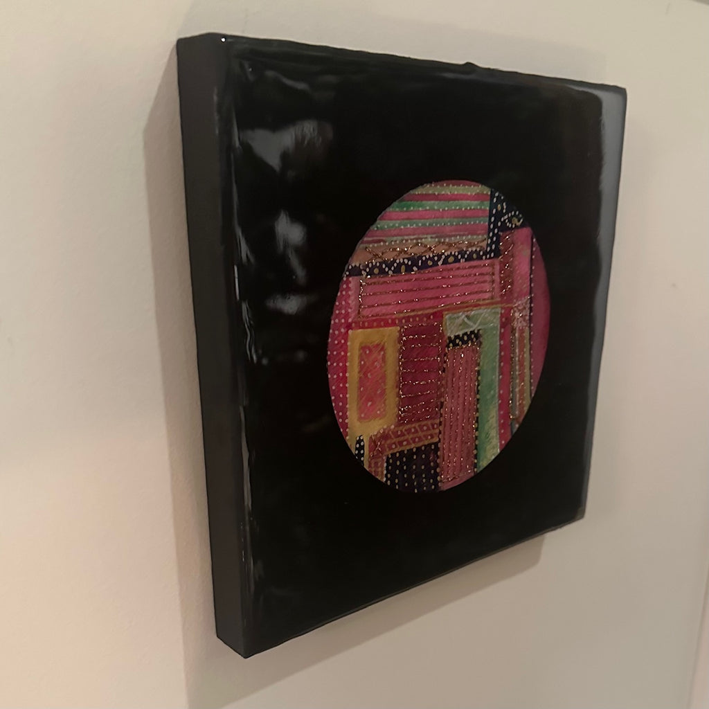 Kvadratisk kunstverk, 20×20 cm, sett fra siden. Blank sort epoxyfinish viser dybden. Fargerikt rundt motiv i rosa, rød og gull med teksturlignende detaljer. Fargesymfoni av Art by Emerense
