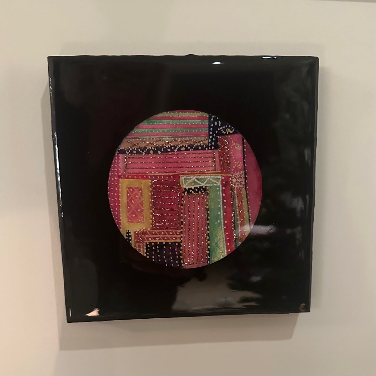 Kvadratisk kunstverk, 20×20 cm, med speilblank sort epoxyoverflate. Rund sirkel i midten med fargerikt tekstilinspirert mønster i rosa, rød, gull, grønn og sort. Fargesymfoni, epoxy og mixed media. Art by Emerense
