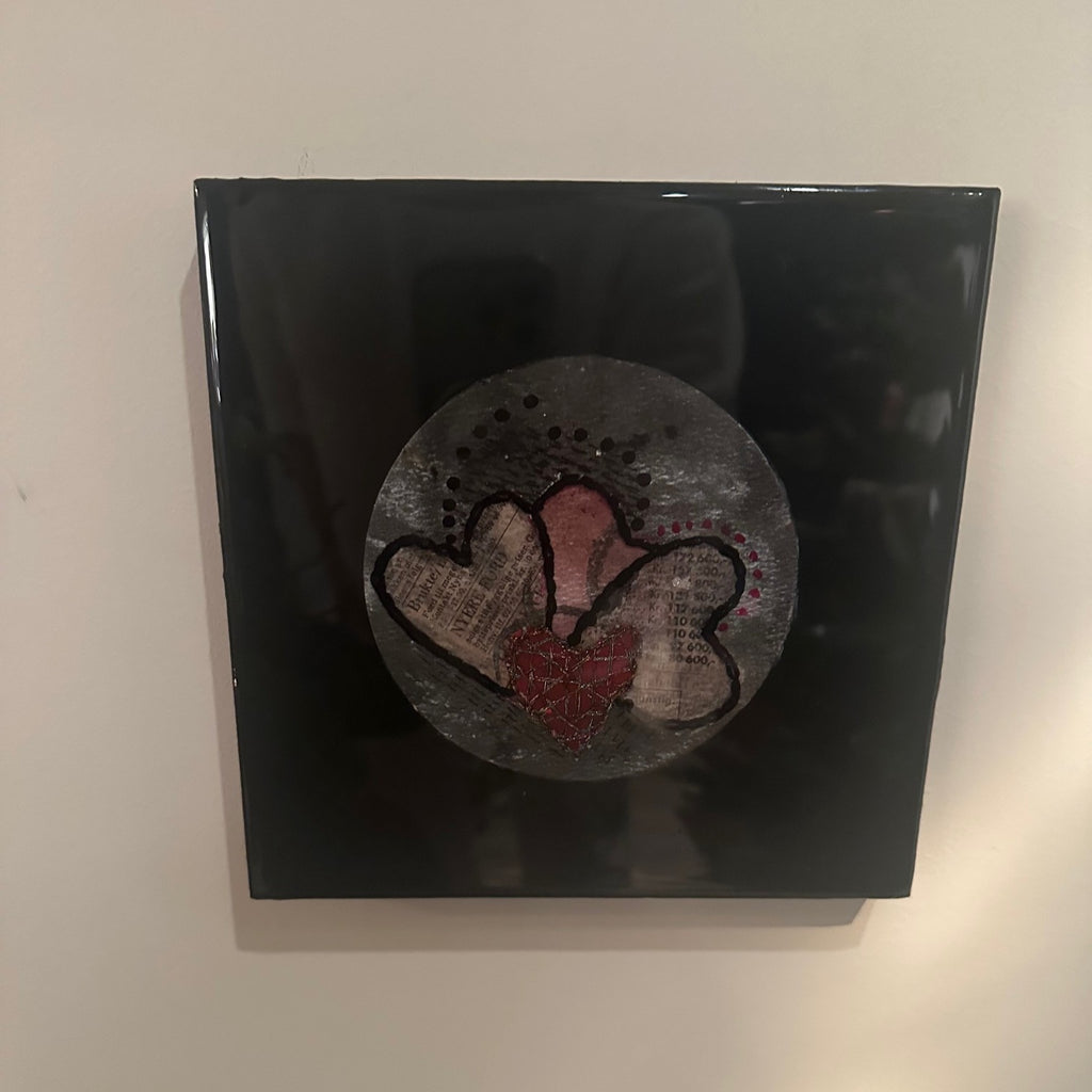Kvadratisk kunstverk, 20×20 cm, med speilblank sort epoxyoverflate. Rund sølvgrå relieffstruktur med tre overlappende hjerter i gammelrosa og rød, brodert med sort tråd og collage-tekstur. Hjertestemme I, epoxy, akvarell og mixed media. Art by Emerense
