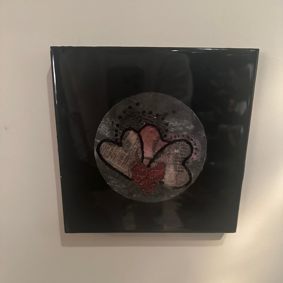Kvadratisk kunstverk, 20×20 cm, med speilblank sort epoxyoverflate. Rund sølvgrå relieffstruktur med tre overlappende hjerter i gammelrosa og rød, brodert med sort tråd og collage-tekstur. Hjertestemme I, epoxy, akvarell og mixed media. Art by Emerense
