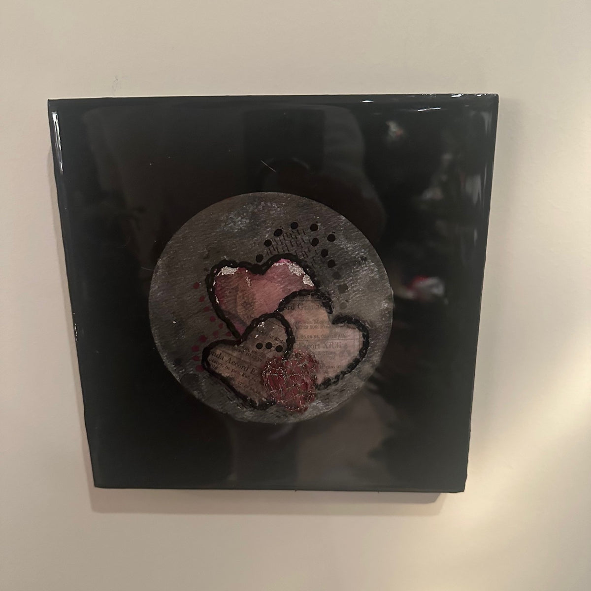 Kvadratisk kunstverk, 20×20 cm, med speilblank sort epoxyoverflate. Rund grå og bronseteksturert sirkel med tre overlappende hjerter i rosa og gammelrosa, brodert med sort tråd. Hjertestemme II, epoxy, akvarell og mixed media. Art by Emerense
