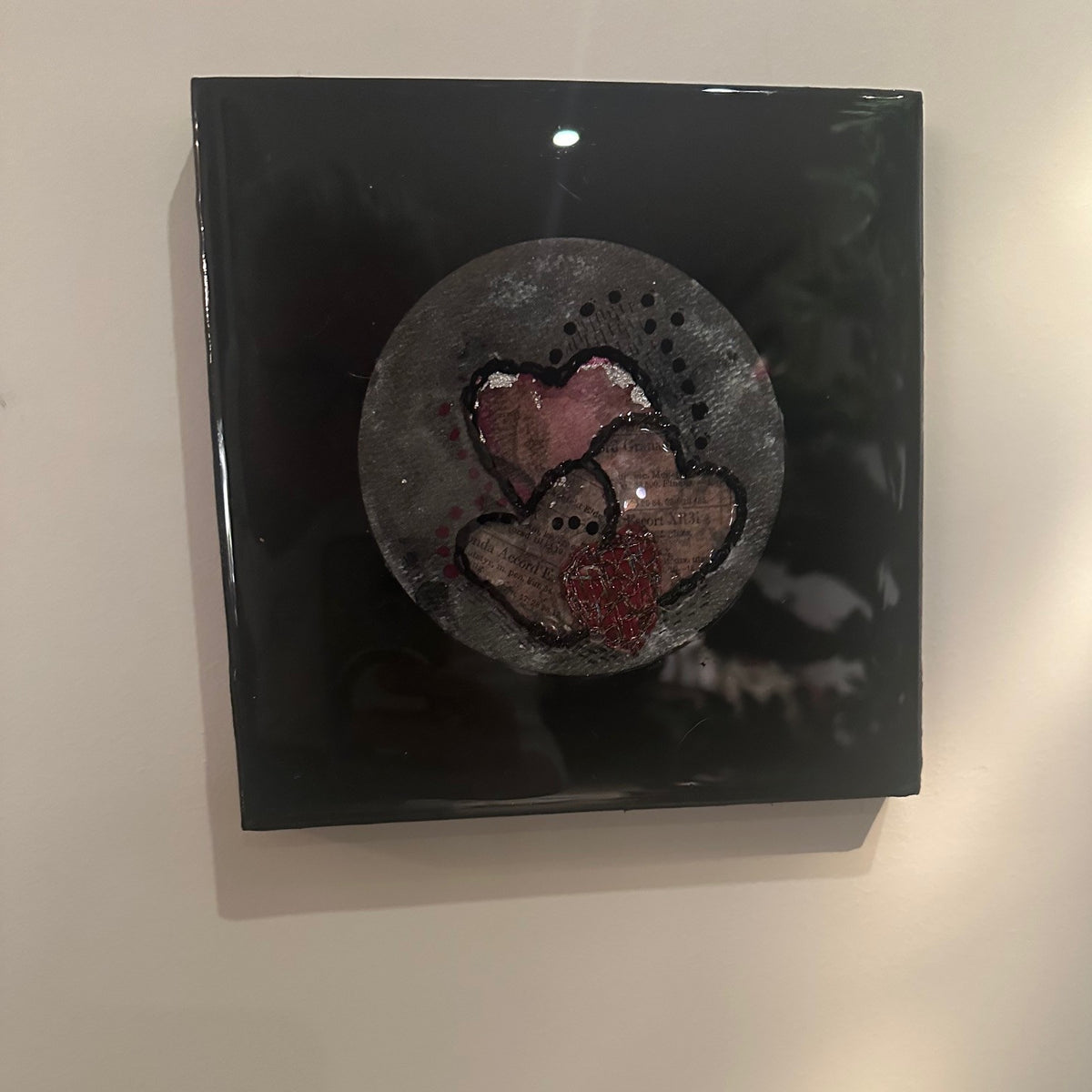 Kvadratisk kunstverk, 20×20 cm, sett ovenfra. Blank sort epoxyfinish med rund grå relieffstruktur og tre rosa hjerter med sort broderi og rød glitrende detalj i midten. Hjertestemme II av Art by Emerense