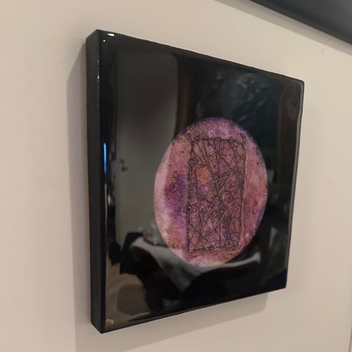 Kvadratisk kunstverk, 20×20 cm, sett fra siden på beige vegg. Tykk blank sort epoxyoverflate reflekterer rommet. Rund lilla og rosa sirkel med mørkt trådenett synlig. Kosmisk vev av Art by Emerense
