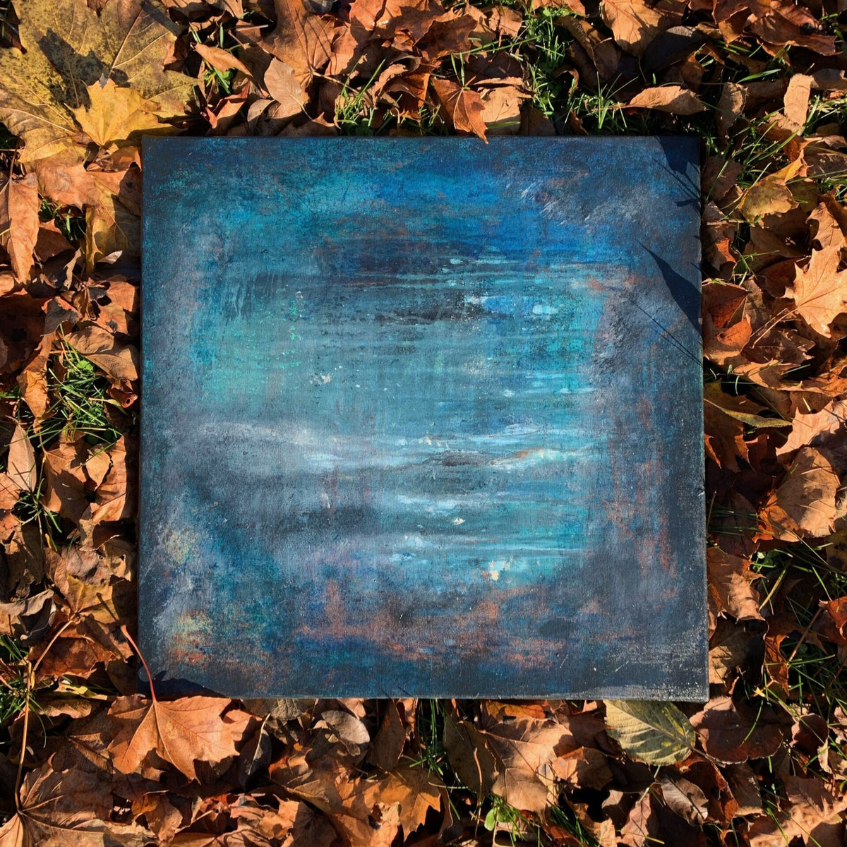Kvadratisk kunstverk, 40×40 cm, med dype blå og turkise nyanser som gjenspeiler vann i skumring. Fotografert på høstløv. Skumring ved Tjønna, akryl og mixmedia. Art by Emerense