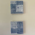 To kvadratiske kunstverk, 20×20 cm, hengt vertikalt på beige vegg. Geometriske blå og sølv felt i mix medium og akryl. Blå firkanter av Art by Emerense