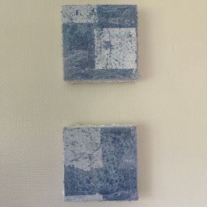 To kvadratiske kunstverk, 20×20 cm, hengt vertikalt på beige vegg. Geometriske blå og sølv felt i mix medium og akryl. Blå firkanter av Art by Emerense