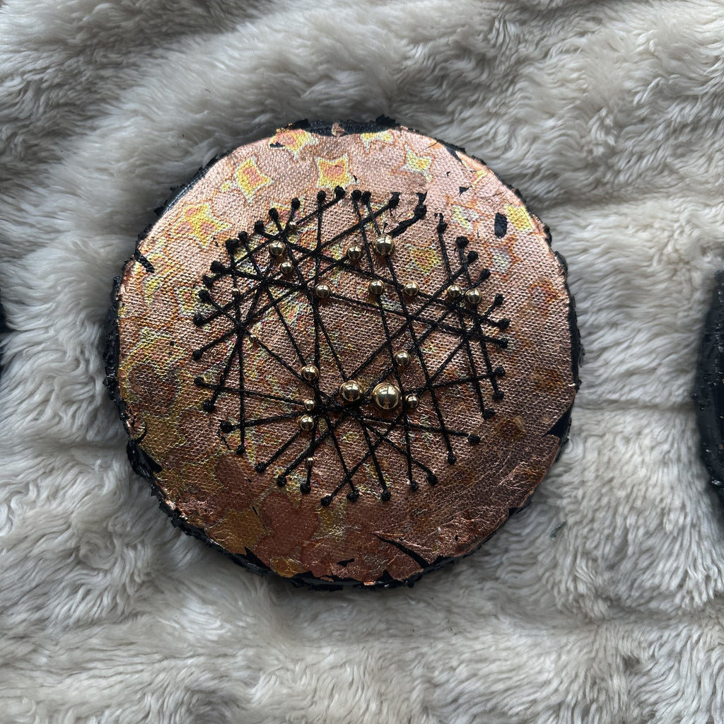 Rundt mixed media-kunstverk, 15 cm diameter, med varm kobber og gullfarget iriserende overflate. Sorte tråder og bronseperler i stjernemønster. Constellations II av Art by Emerense