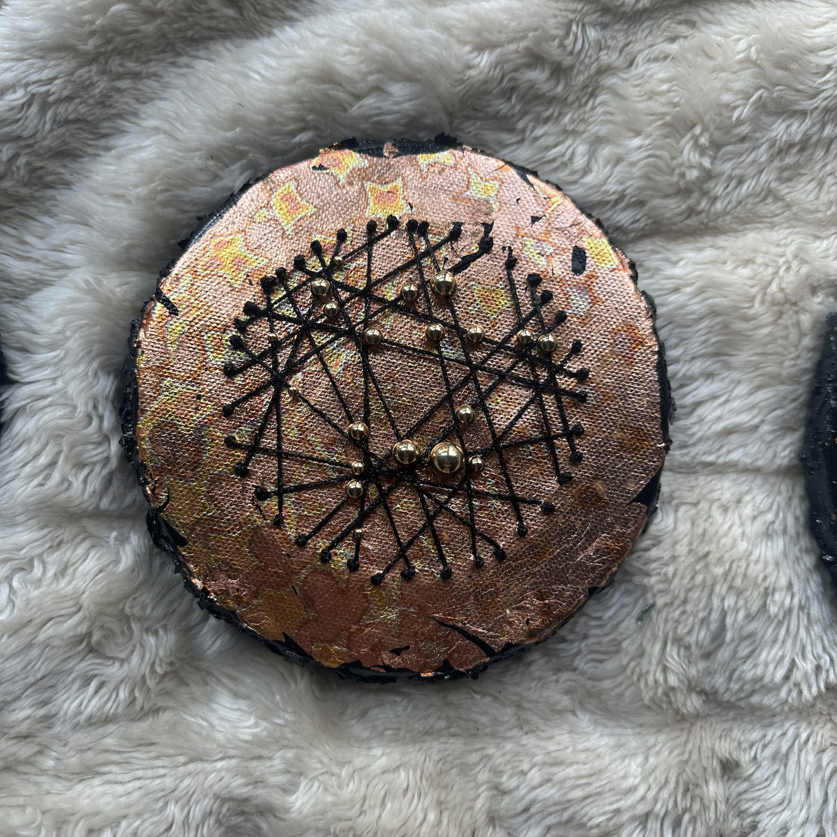 Rundt mixed media-kunstverk, 15 cm diameter, med varm kobber og gullfarget iriserende overflate. Sorte tråder og bronseperler i stjernemønster. Constellations II av Art by Emerense