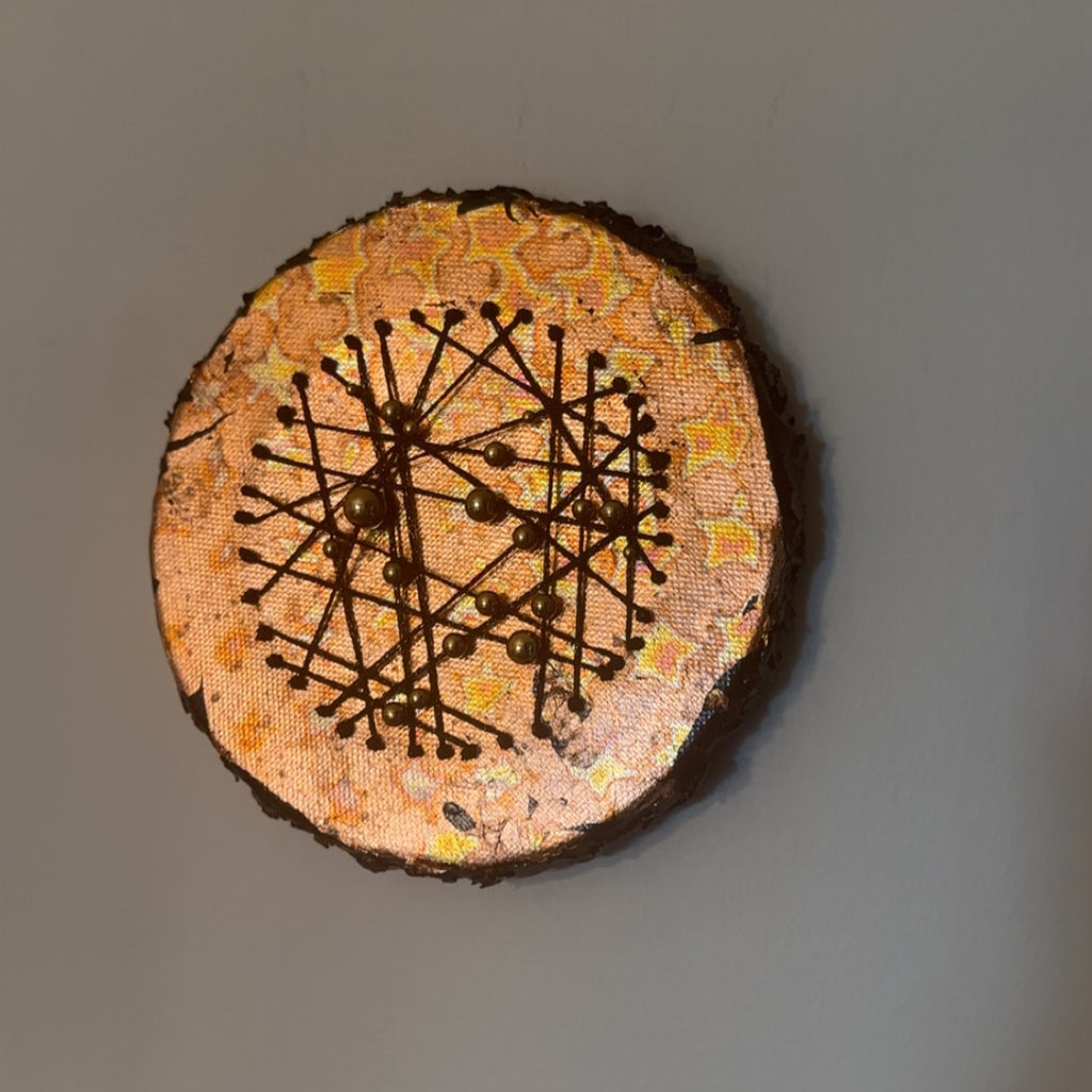 Rundt mixed media-kunstverk, 15 cm diameter, hengt på grå vegg. Varm kobber og oransje overflate med sorte tråder og bronseperler. Constellations II av Art by Emerense