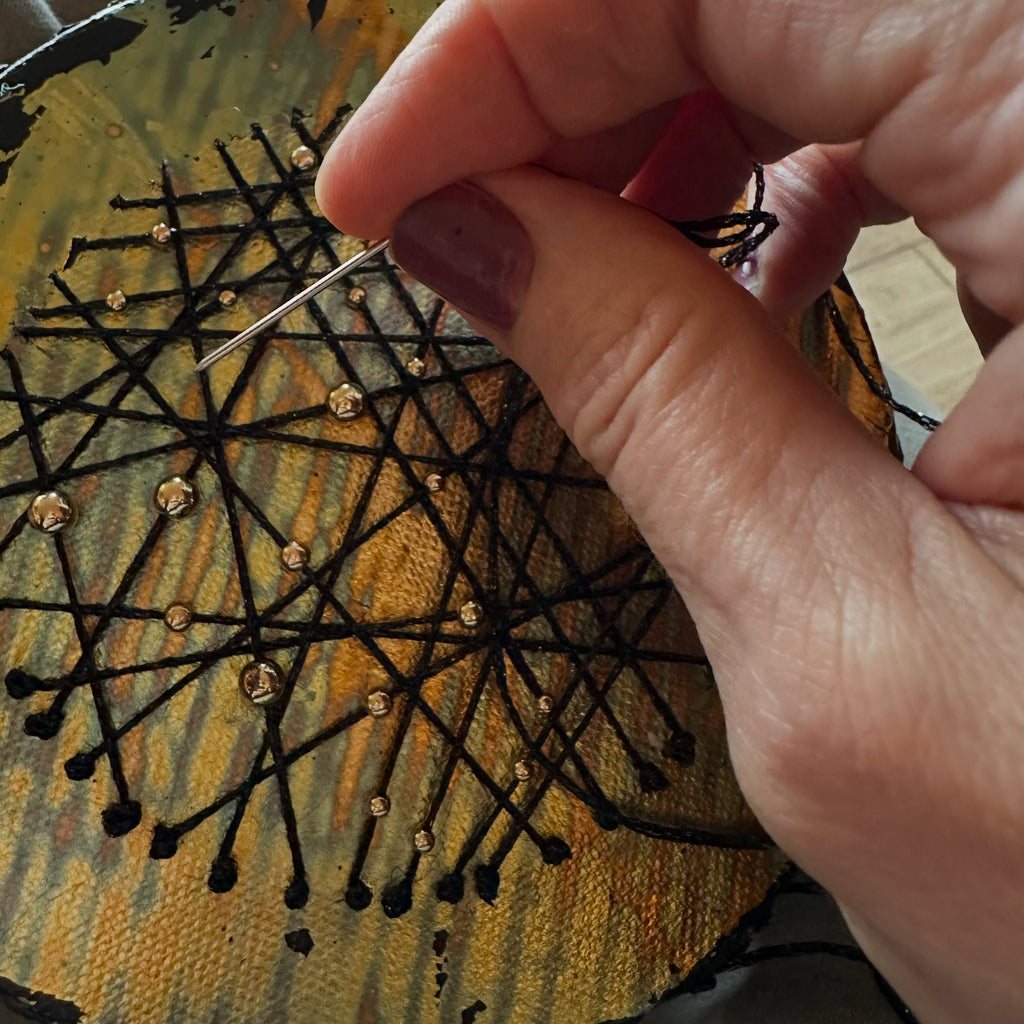 Nærbilde av kunstnerens hånd som syr svarte tråder med nål på et rundt lerret i metalliske gulltoner med små gullfargede detaljer. Arbeid med et kunstverk i Constellations-serien.