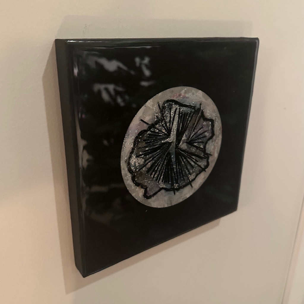 Kvadratisk kunstverk, 20×20 cm, sett fra siden. Tykk blank sort epoxyfinish viser dybden. Rund sølvgrå struktur med sort abstrakt relieff i midten. Nattens symfoni av Art by Emerense
