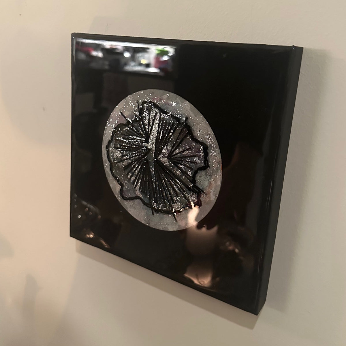 Kvadratisk kunstverk, 20×20 cm, sett fra siden med lys som reflekteres i blank sort epoxyoverflate. Rund sølvglitter sirkel med sort abstrakt relieff synlig. Nattens symfoni av Art by Emerense