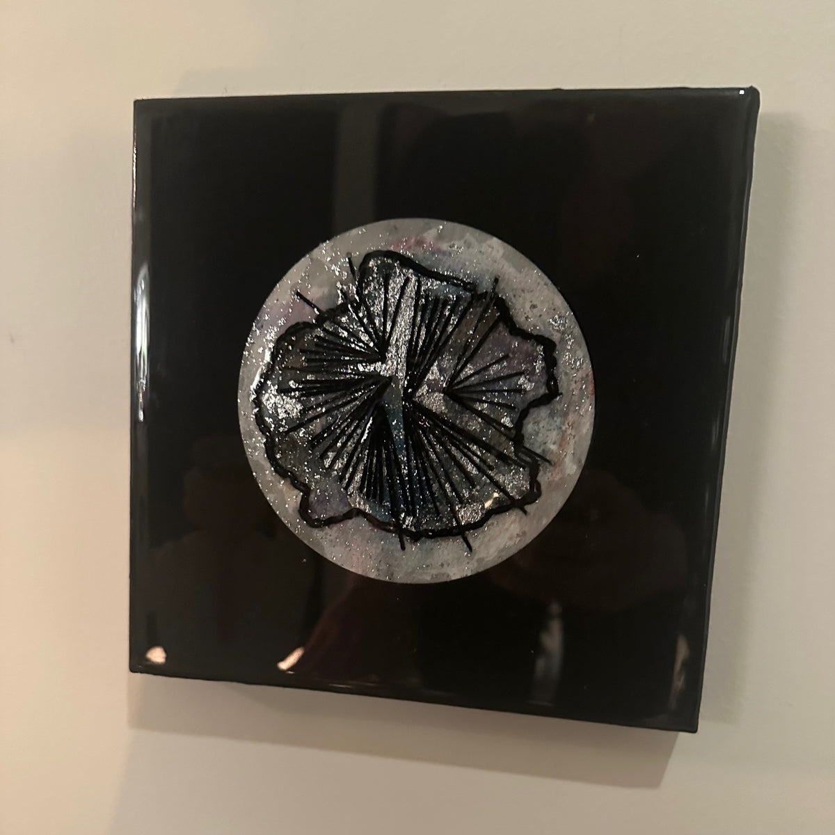 Kvadratisk kunstverk, 20×20 cm, med speilblank sort epoxyoverflate. Rund sølv og grå sirkul struktur i midten med abstrakt sort blomsterlignende relieff. Nattens symfoni, epoxy og mixed media. Art by Emerense