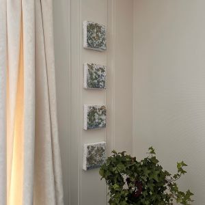 Fire kvadratiske kunstverk hengt vertikalt på hvit vegg ved vindu med plante i forgrunnen. Vinterstemning, mix medium. Art by Emerense